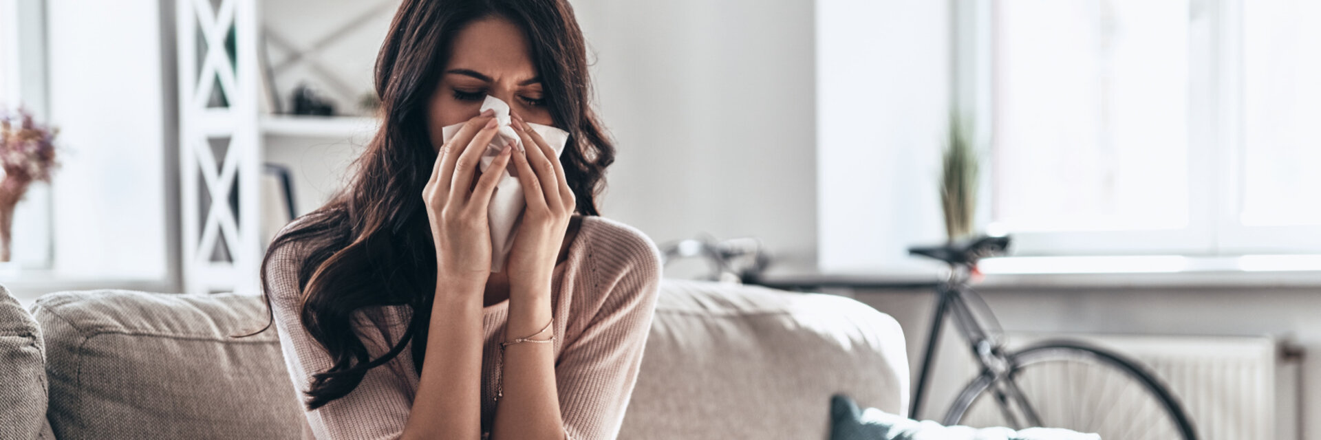 Cold and flu. Sick young woman blowing the nose using tissue paper while sitting on the sofa at home, Woman at home with flu or allergy symptoms cleaning her nose
