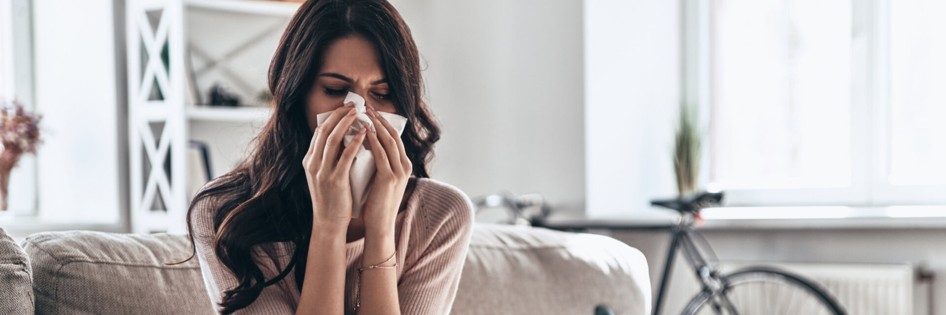 Cold and flu. Sick young woman blowing the nose using tissue paper while sitting on the sofa at home, Woman at home with flu or allergy symptoms cleaning her nose
