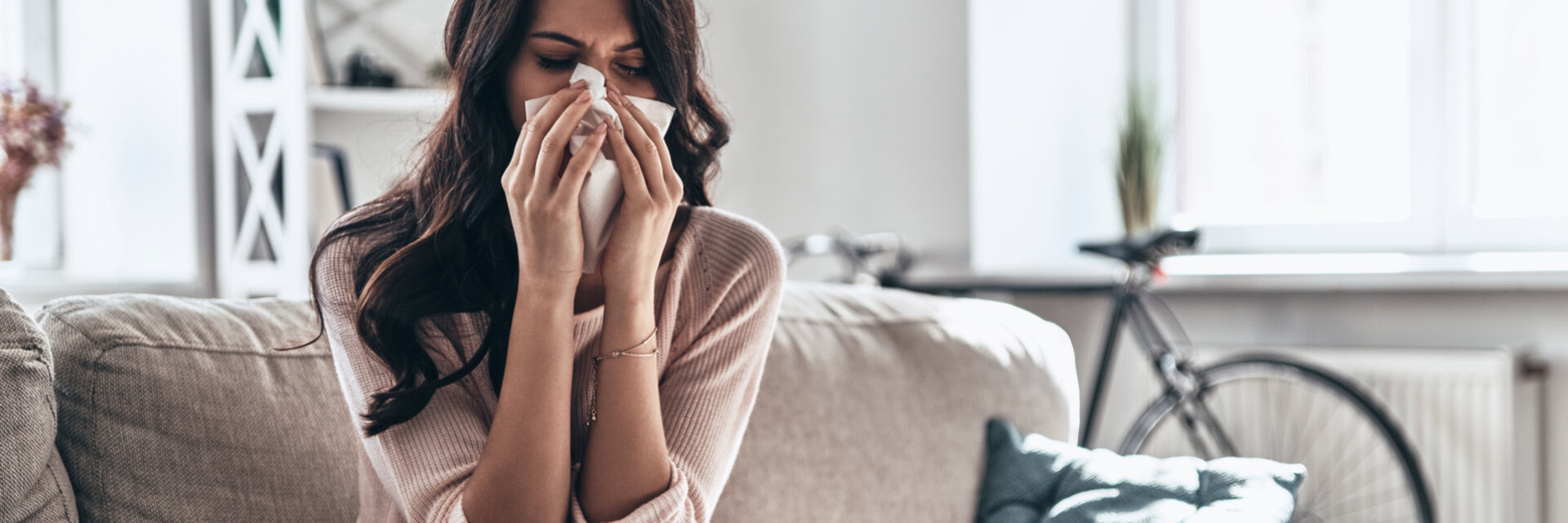 Cold and flu. Sick young woman blowing the nose using tissue paper while sitting on the sofa at home, Woman at home with flu or allergy symptoms cleaning her nose
